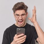 Pissed off annoyed man screams angrily keeps palm raised stares at smartphone being outraged after rough conversation wears round spectacles casual black t shirt isolated over white background